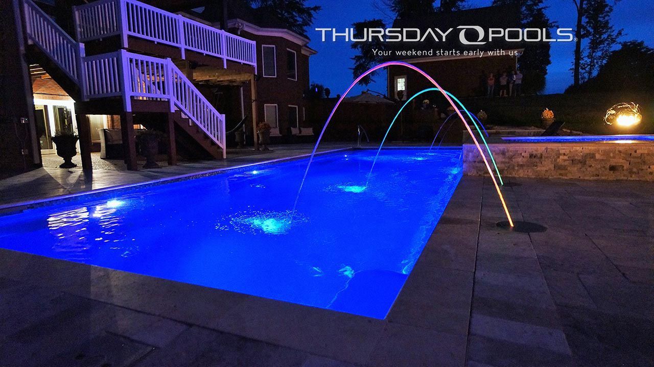 Nighttime backyard pool glowing blue with arcing water jets and a lit deck beside a house