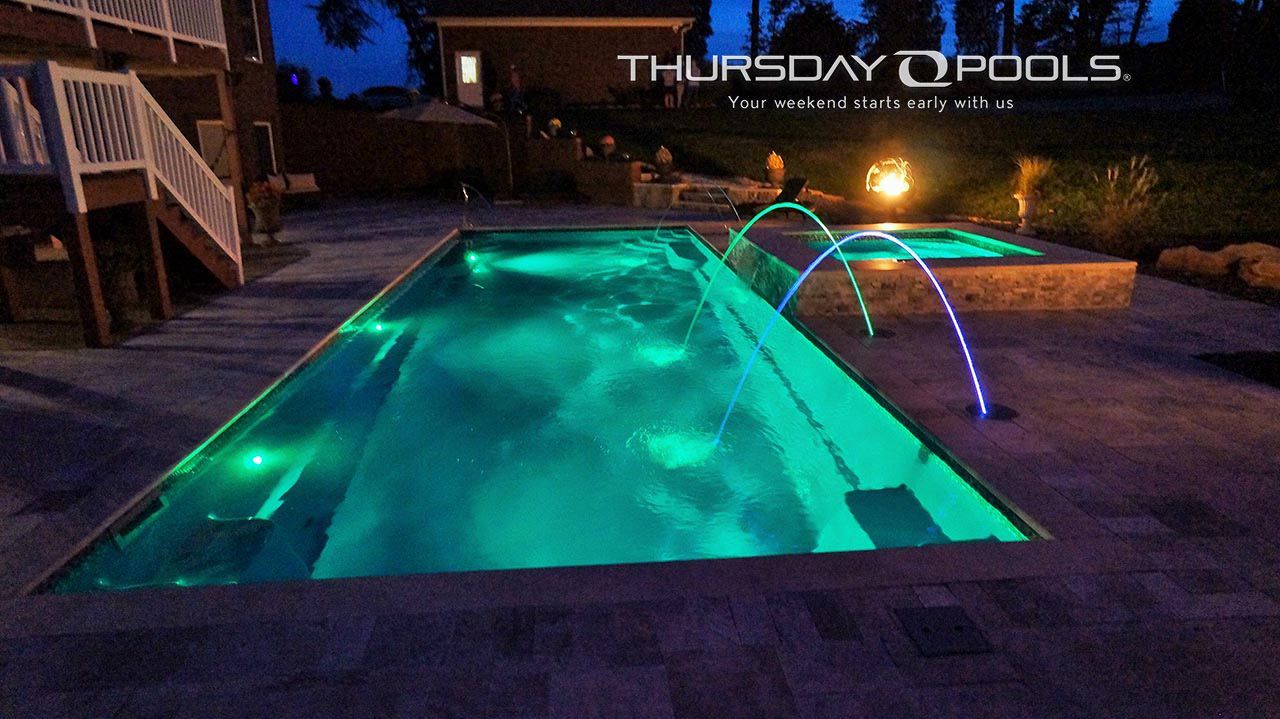Backyard swimming pool glowing turquoise at night with an arched water feature and deck lighting