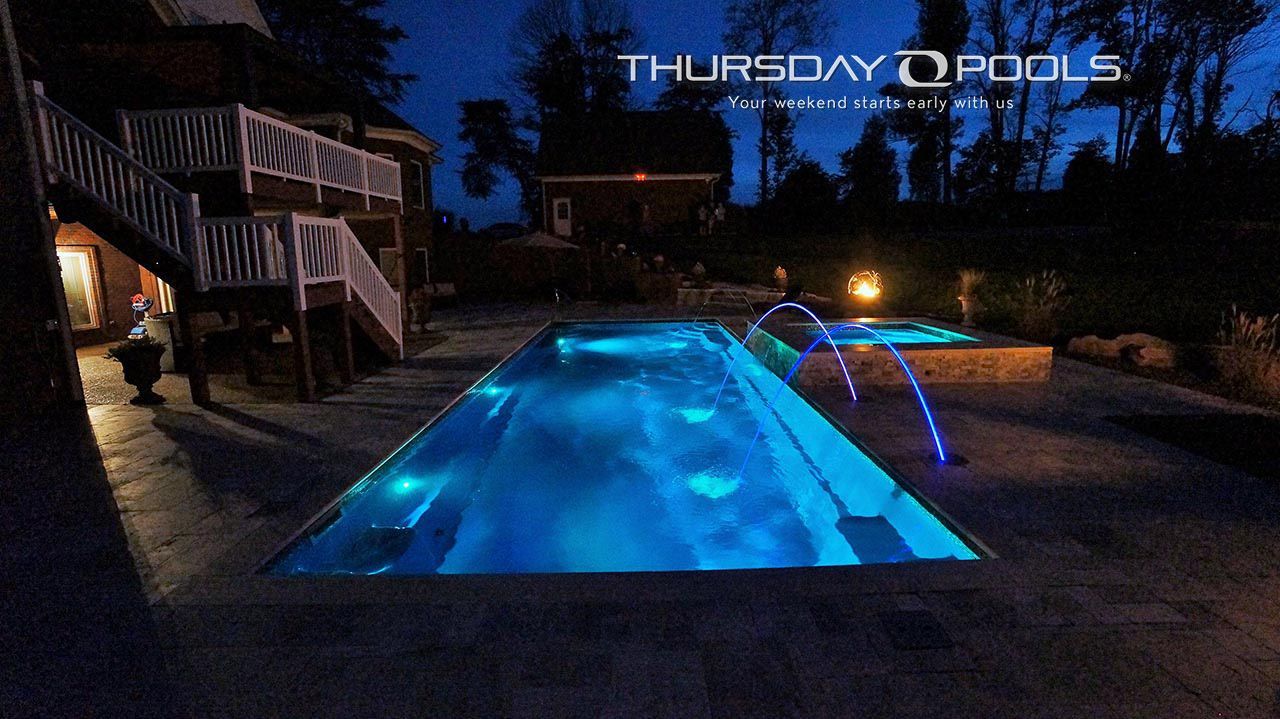Backyard swimming pool glowing blue at night with patio lights and house deck in the background