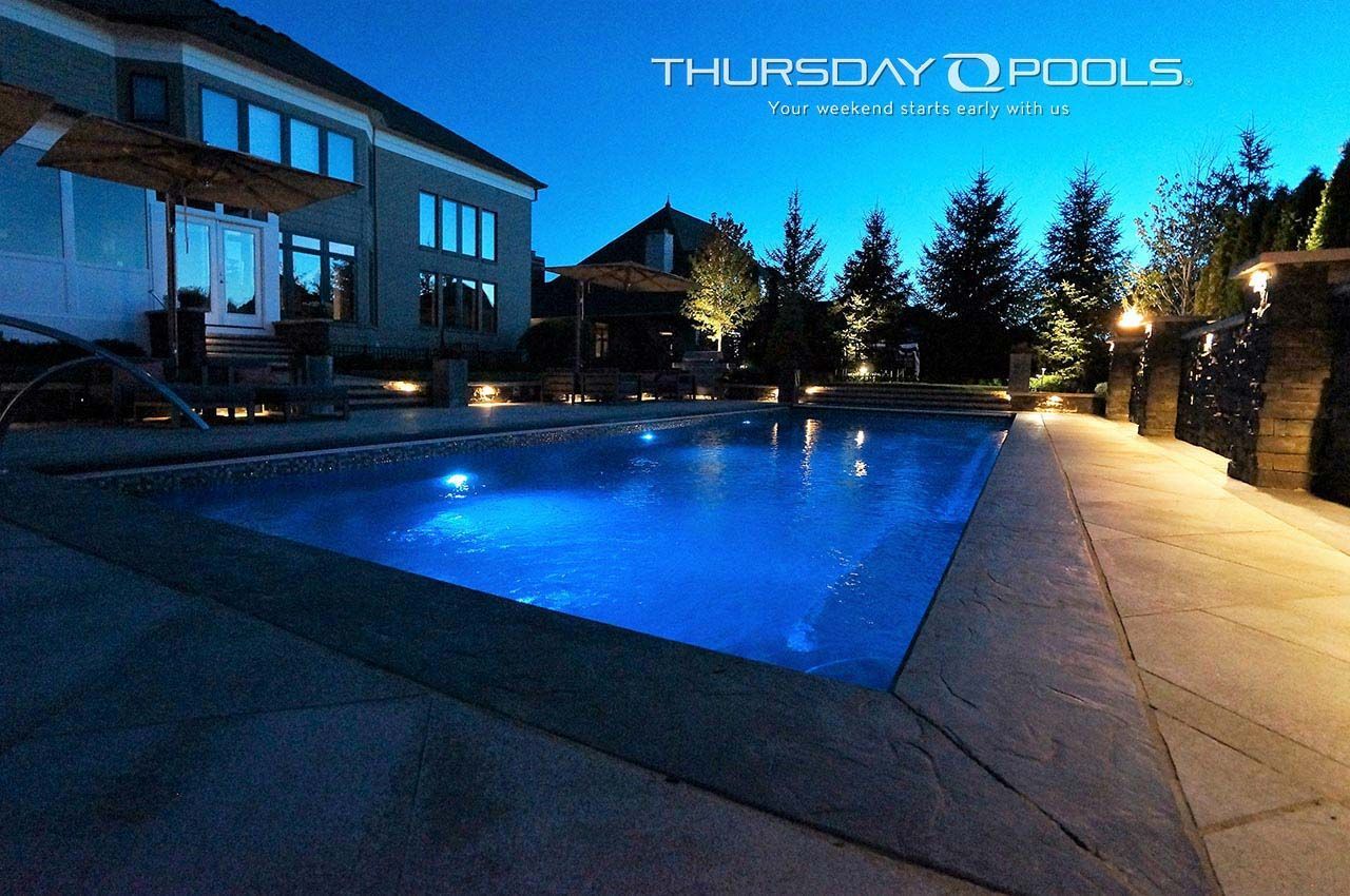 Blue-lit backyard swimming pool at dusk beside a house, with lights and trees in the background