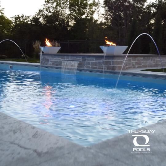 Backyard pool at dusk with glowing fire bowls and arcing water jets