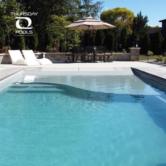 Backyard swimming pool with lounge chairs, umbrella, and stone patio under a clear sky