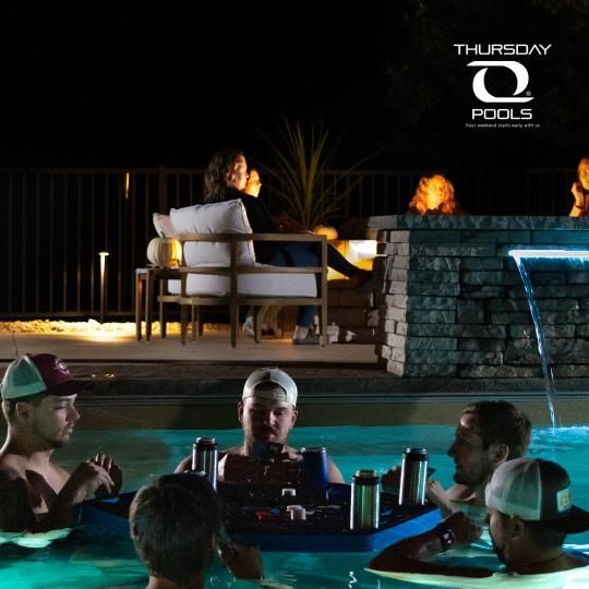 People relaxing in a pool at night beside a lit patio and fountain