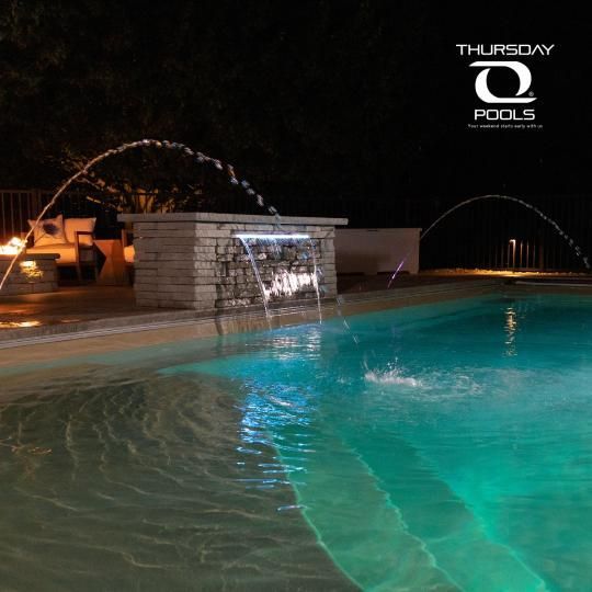 Nighttime swimming pool with turquoise water and a lit waterfall feature, branded Thursday Pools.