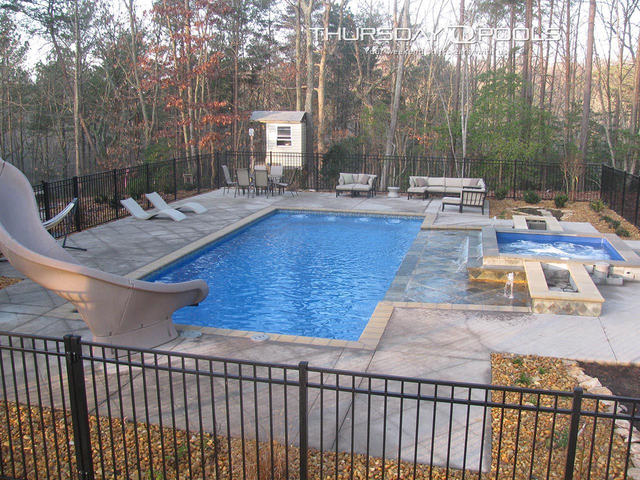 Backyard pool with blue water, slide, lounge chairs, and a hot tub surrounded by trees