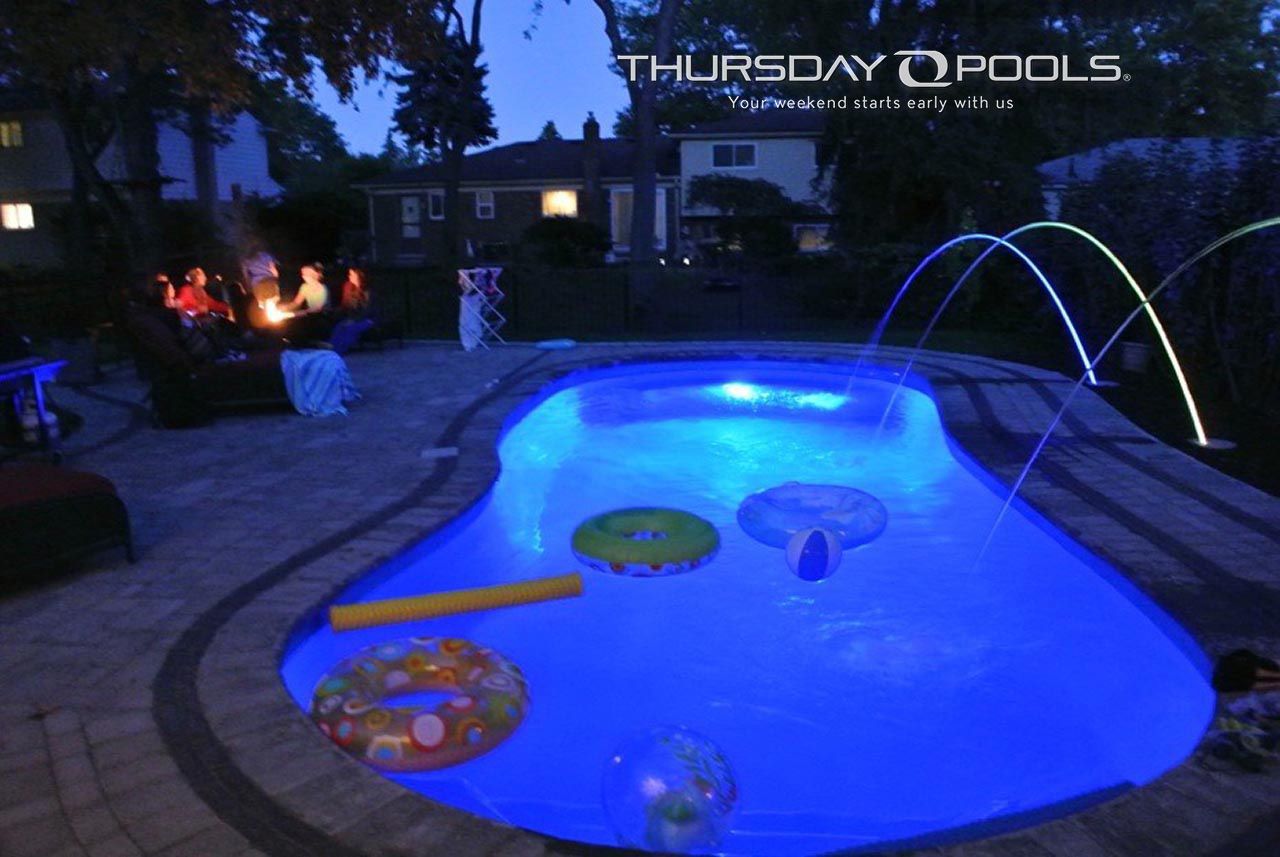 Backyard swimming pool glowing blue at night, with pool floats and a small arch fountain.
