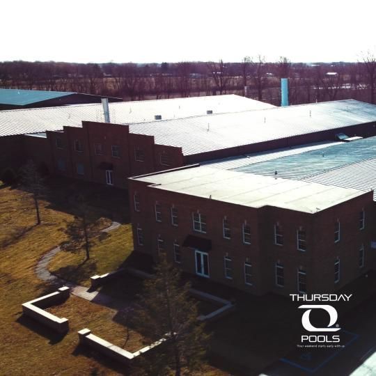 Aerial view of a brick building with a white roof in a wooded area under a bright sky.