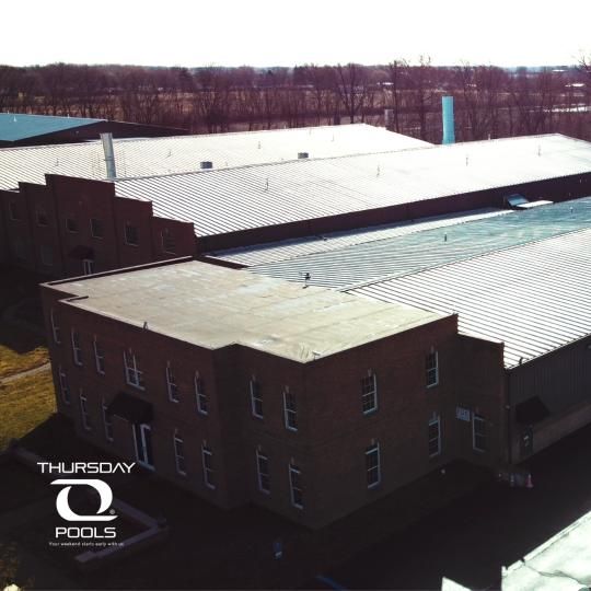 Aerial view of a brick industrial building with a white metal roof and surrounding trees under bright sunlight