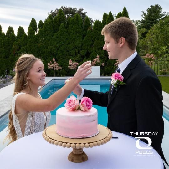 Bride feeds groom cake at an outdoor wedding reception beside a pink cake and blue pool.