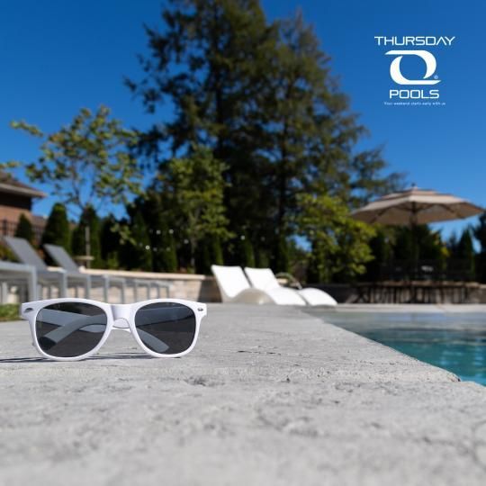 White sunglasses by a sunny pool with blue water, lounge chairs, and trees in the background.