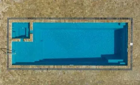 Overhead view of rectangular swimming pool with attached square area, surrounded by light brown paving.