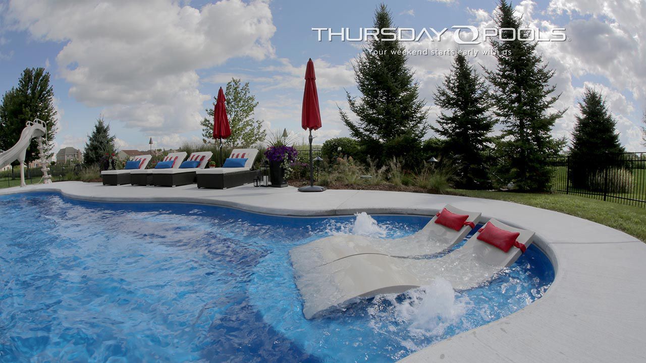 Outdoor pool with blue water, white lounge chairs, red umbrellas, and evergreen trees under cloudy sky