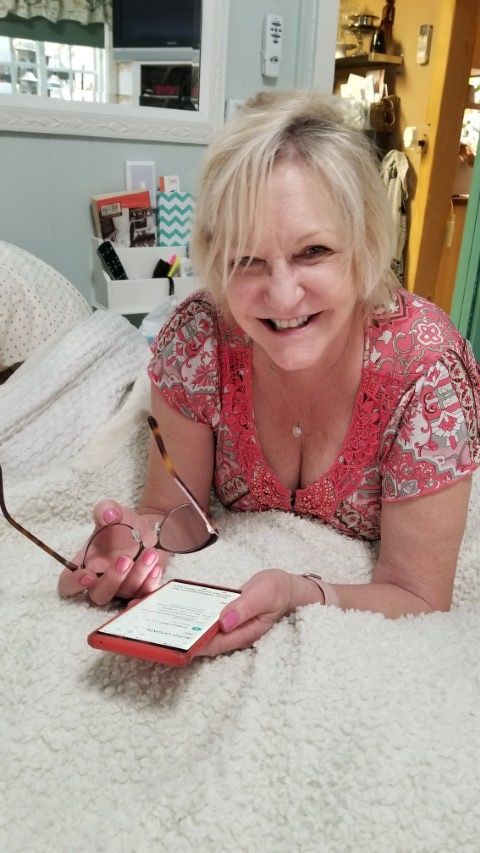 Bexx is lying on a bed holding a cell phone and smiling.