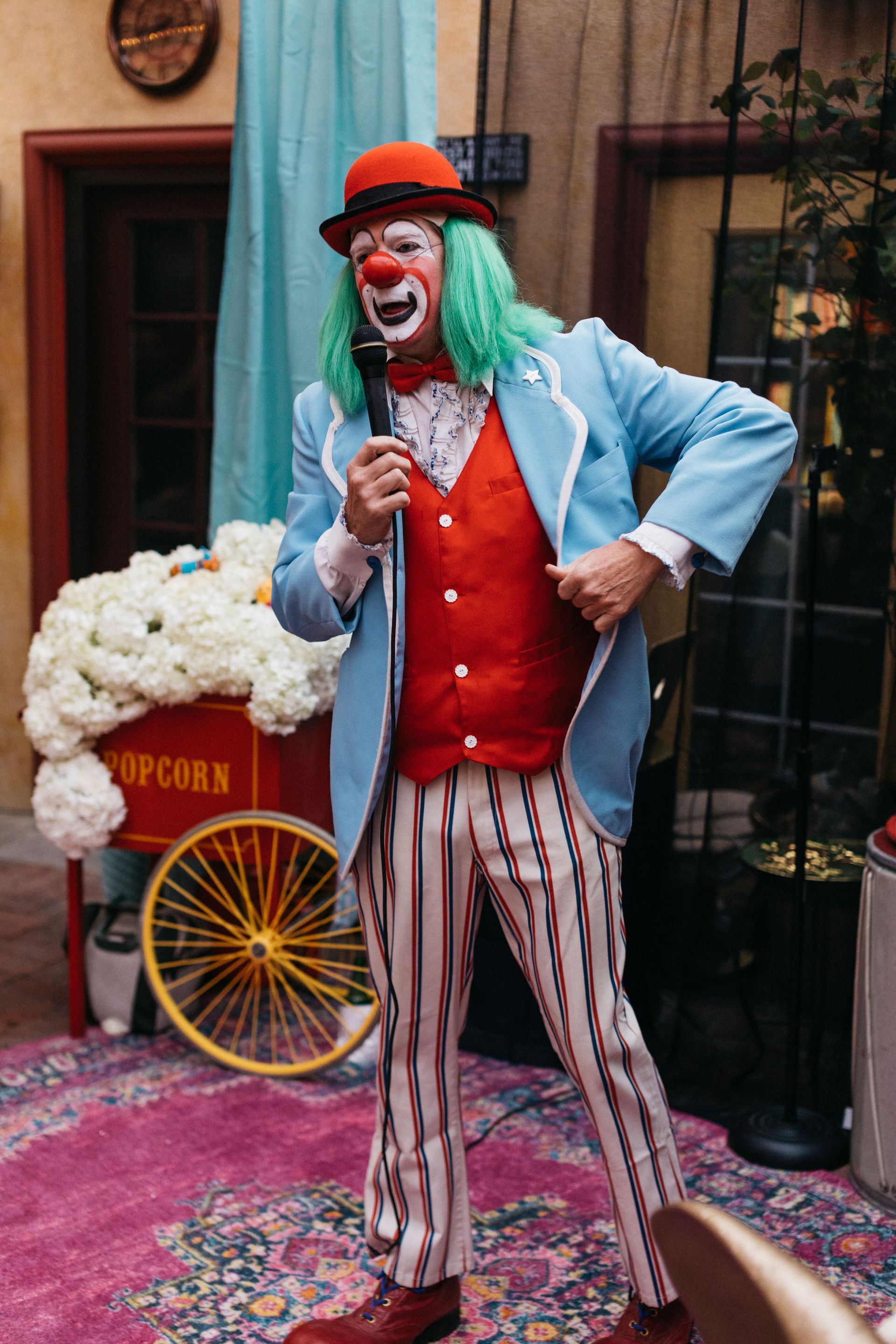 A clown is holding a microphone in front of a popcorn cart.