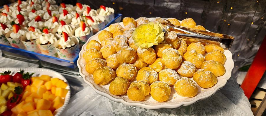 A plate of doughnuts with powdered sugar on top is on a table with other desserts.