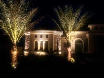 A house is lit up at night with palm trees in front of it.