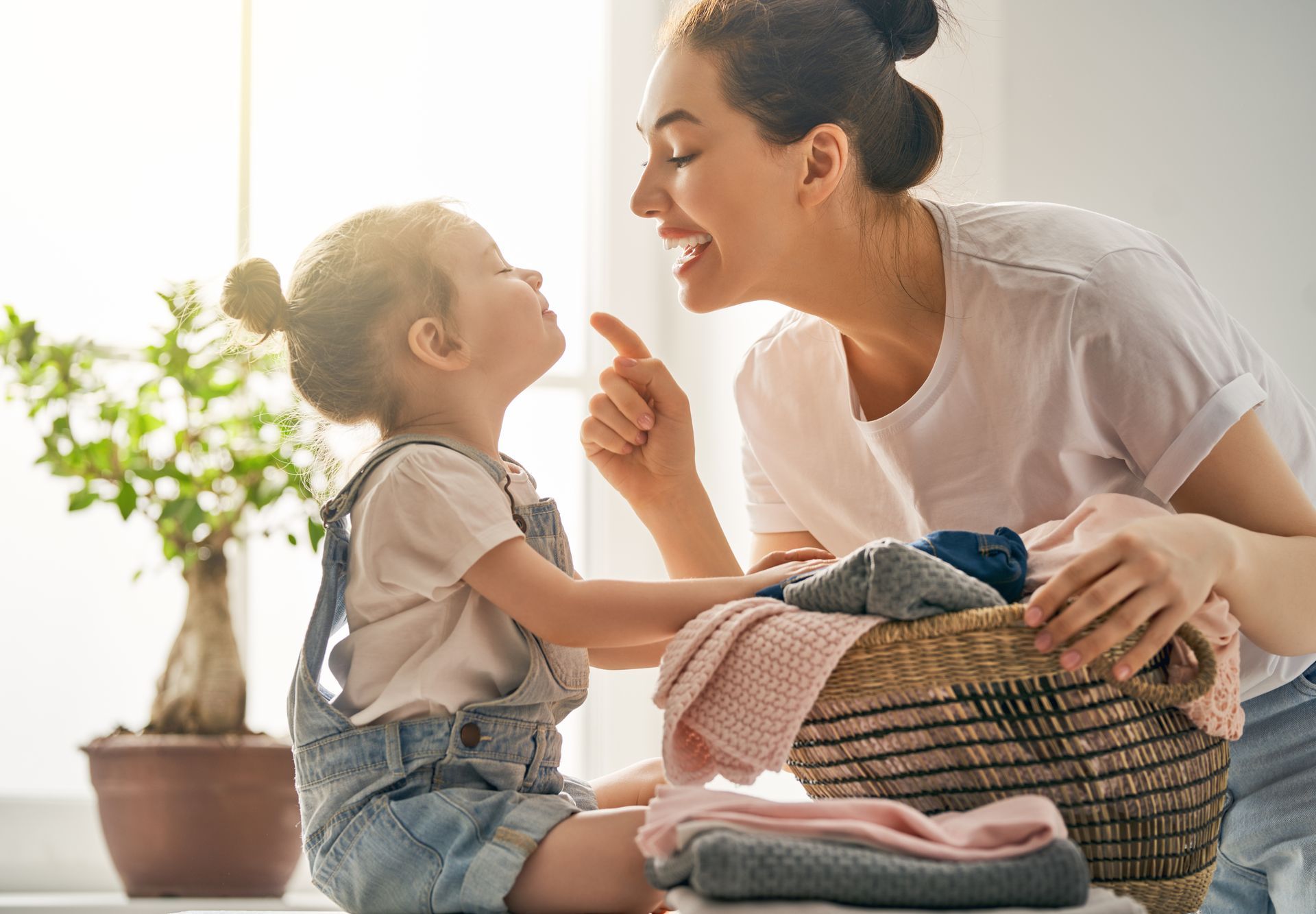 Woman smiling at toddler beside a basket of folded laundry in a sunlit room
