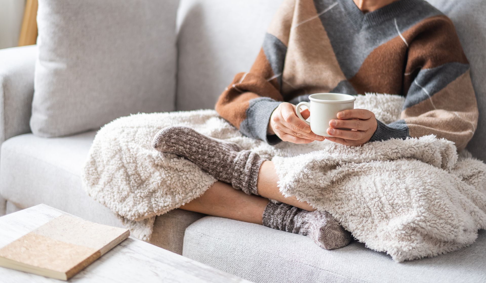 Person wrapped in a cozy blanket on a sofa, holding a mug beside a laptop
