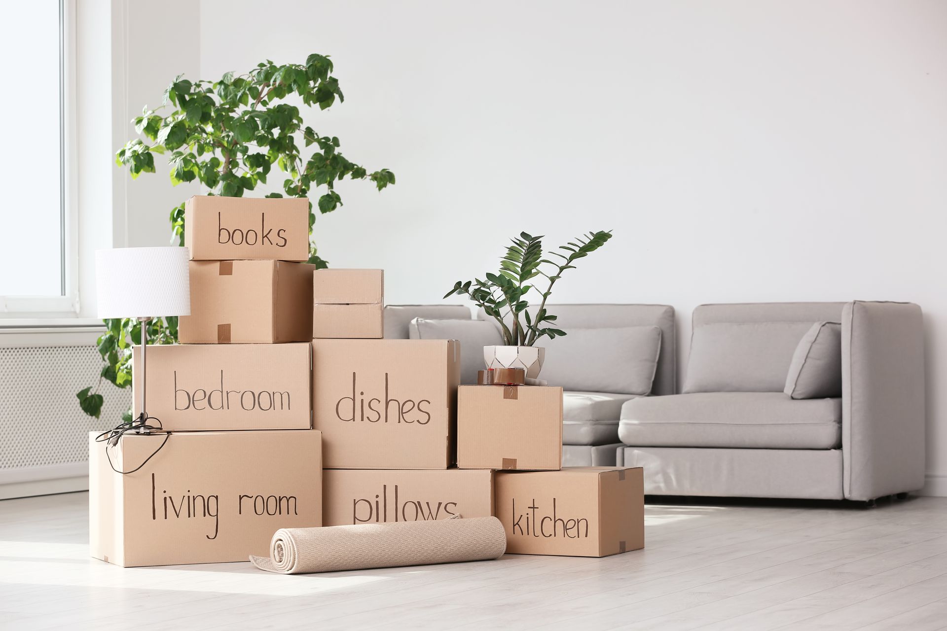 Cardboard moving boxes labeled with room names beside a beige sofa in a bright living room