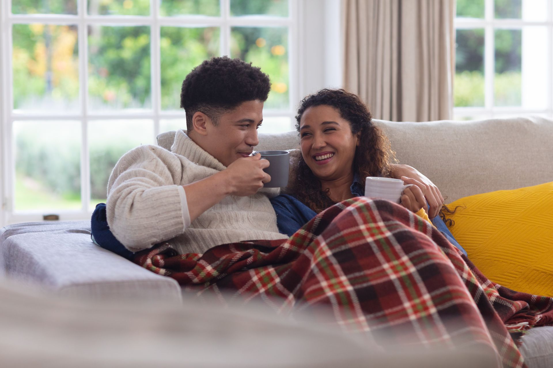 Couple cuddling on a couch under a plaid blanket, smiling with mugs in a bright living room.