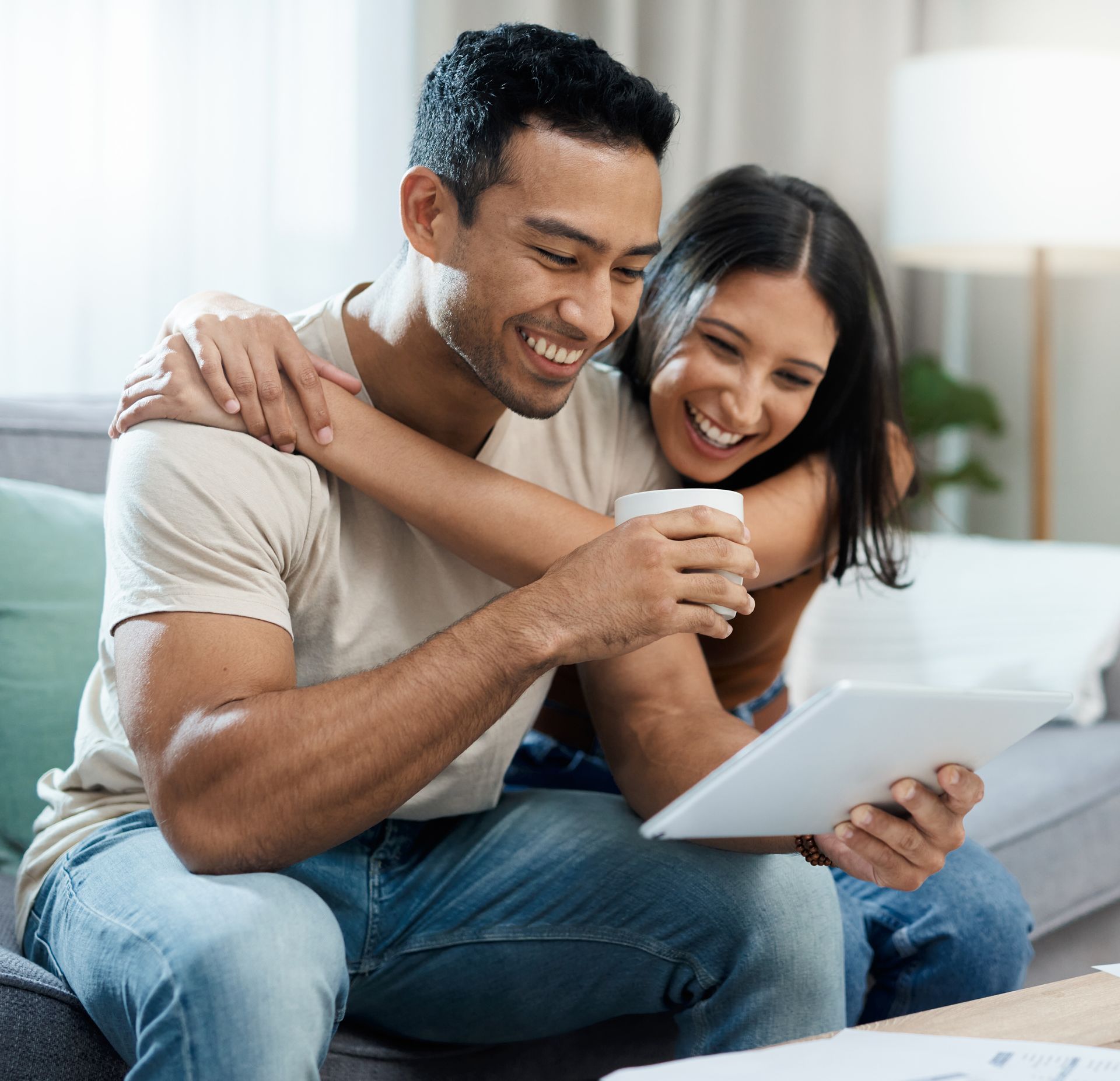 Couple smiling on a couch, looking at a tablet and holding a cup in a bright living room