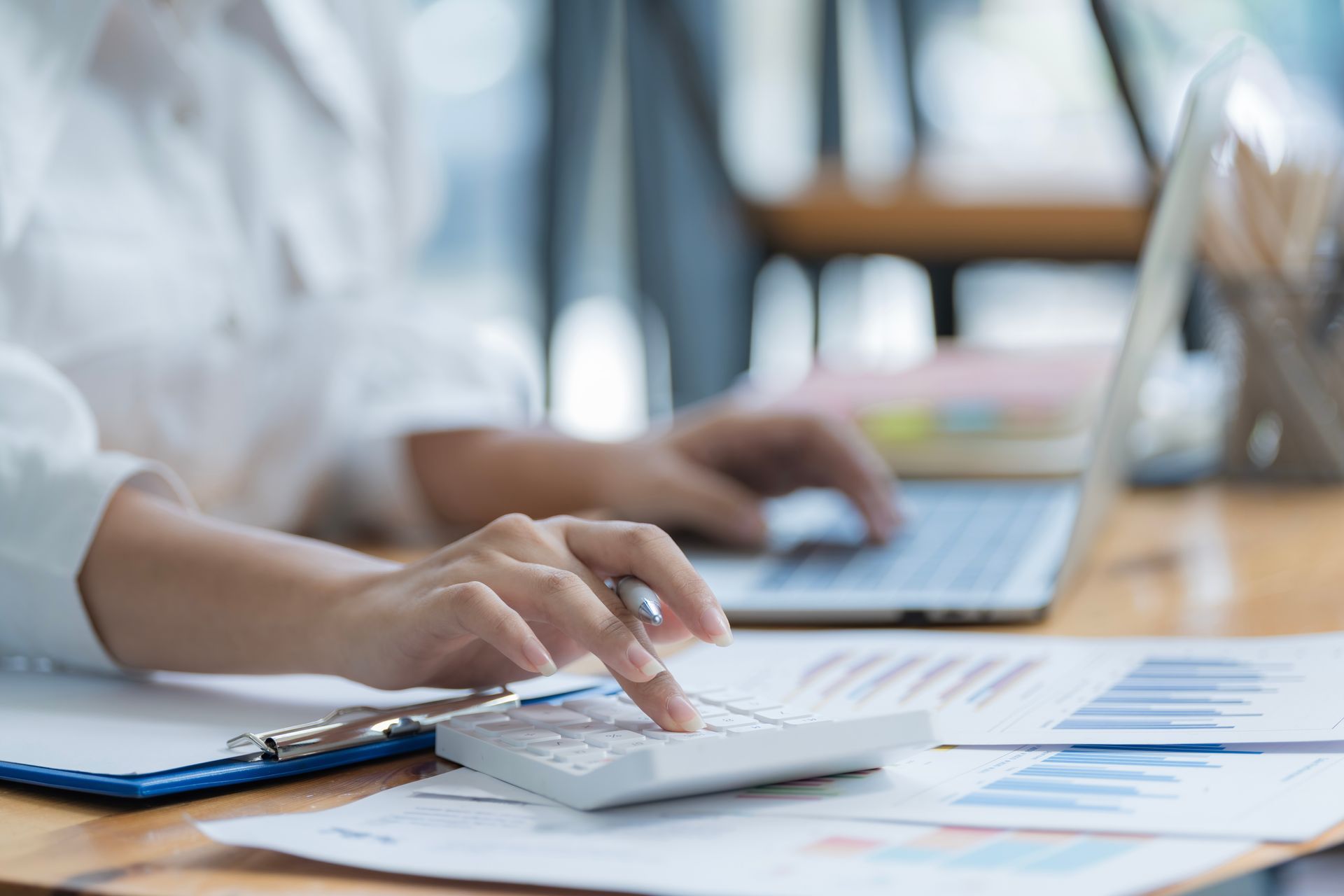 Hands using a calculator beside a laptop and paperwork on a desk