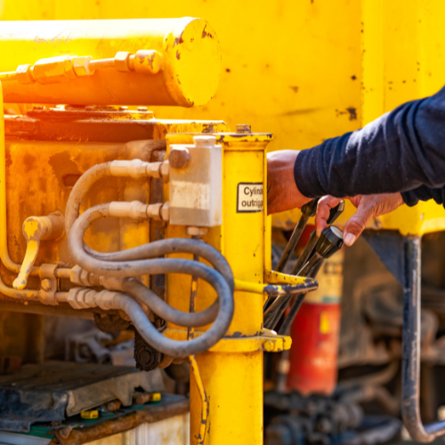 Person operating yellow machinery, possibly construction equipment. Controls and hydraulic lines are visible.