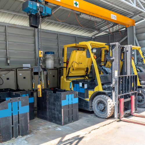 Yellow forklift and overhead crane in a warehouse next to stacked containers and equipment.