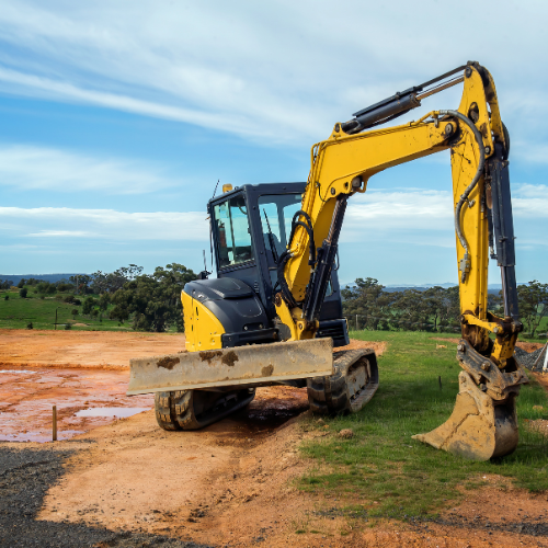 Yellow excavator on a construction site with a green grassy area, under a partly cloudy blue sky.