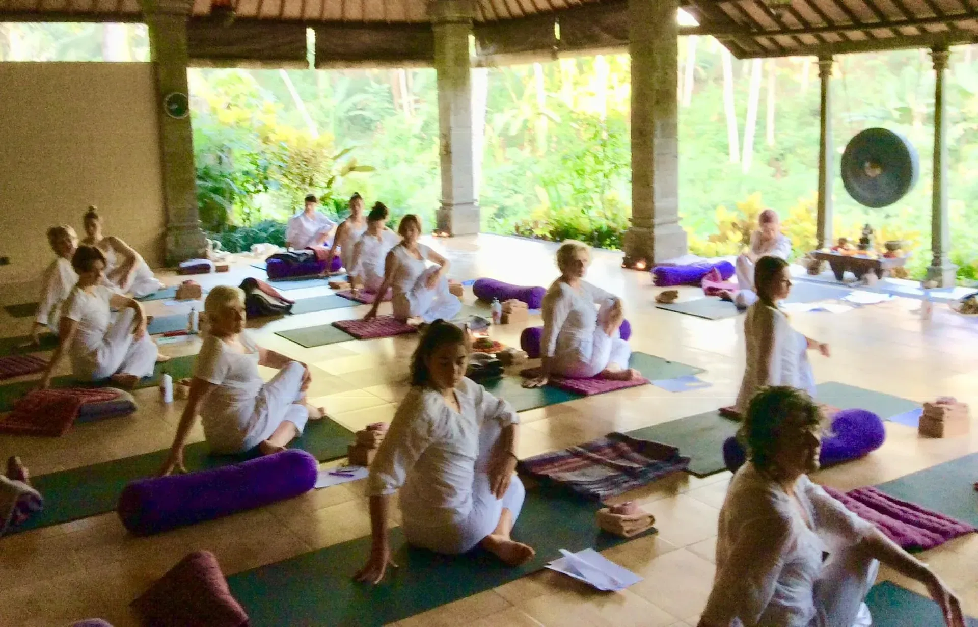 A group in white clothing practices yoga on mats inside an open-air pavilion surrounded by lush tropical greenery.