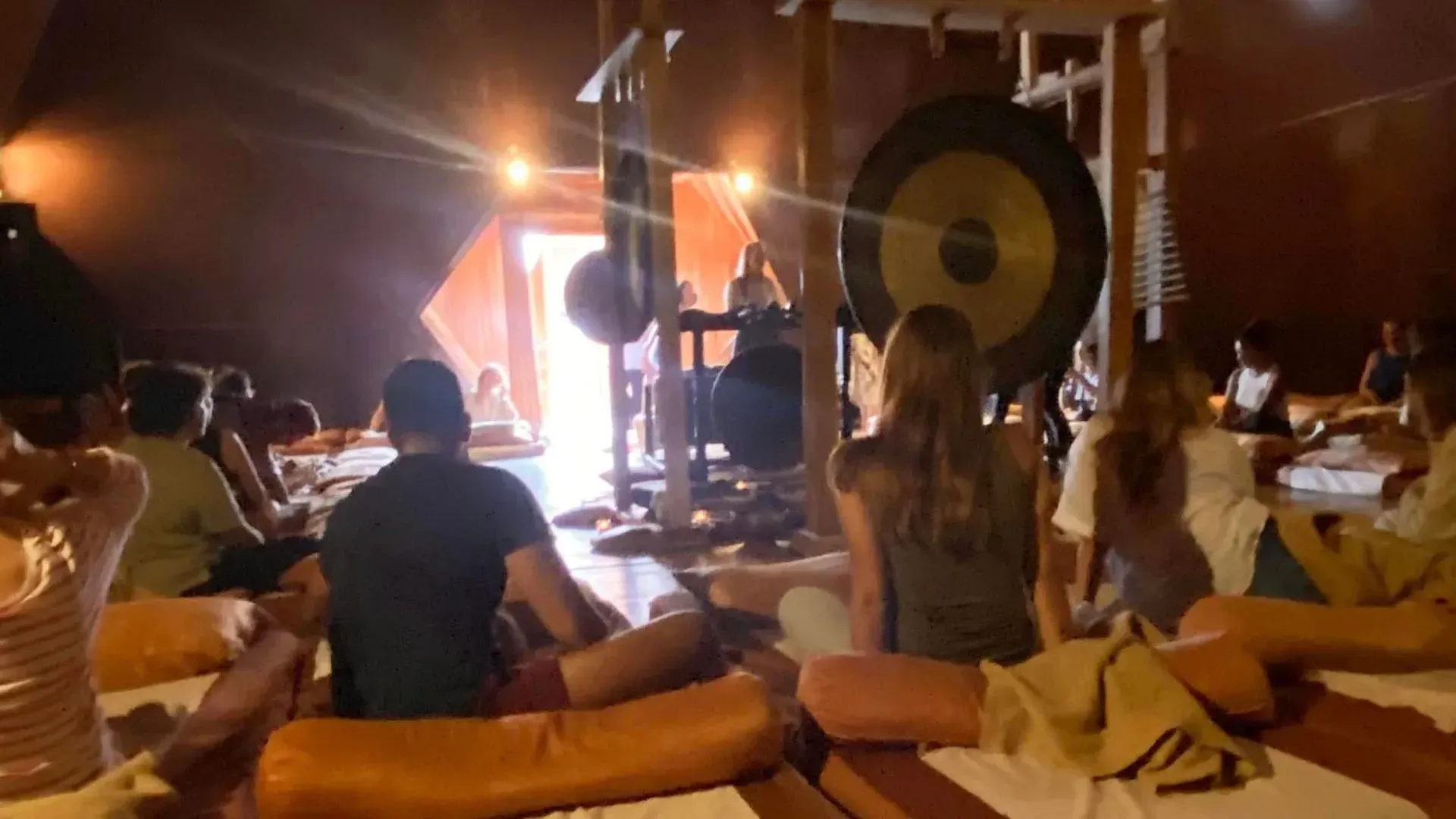 People sitting on mats in a dimly lit, warm-toned room facing a large gong stand during a sound meditation session.