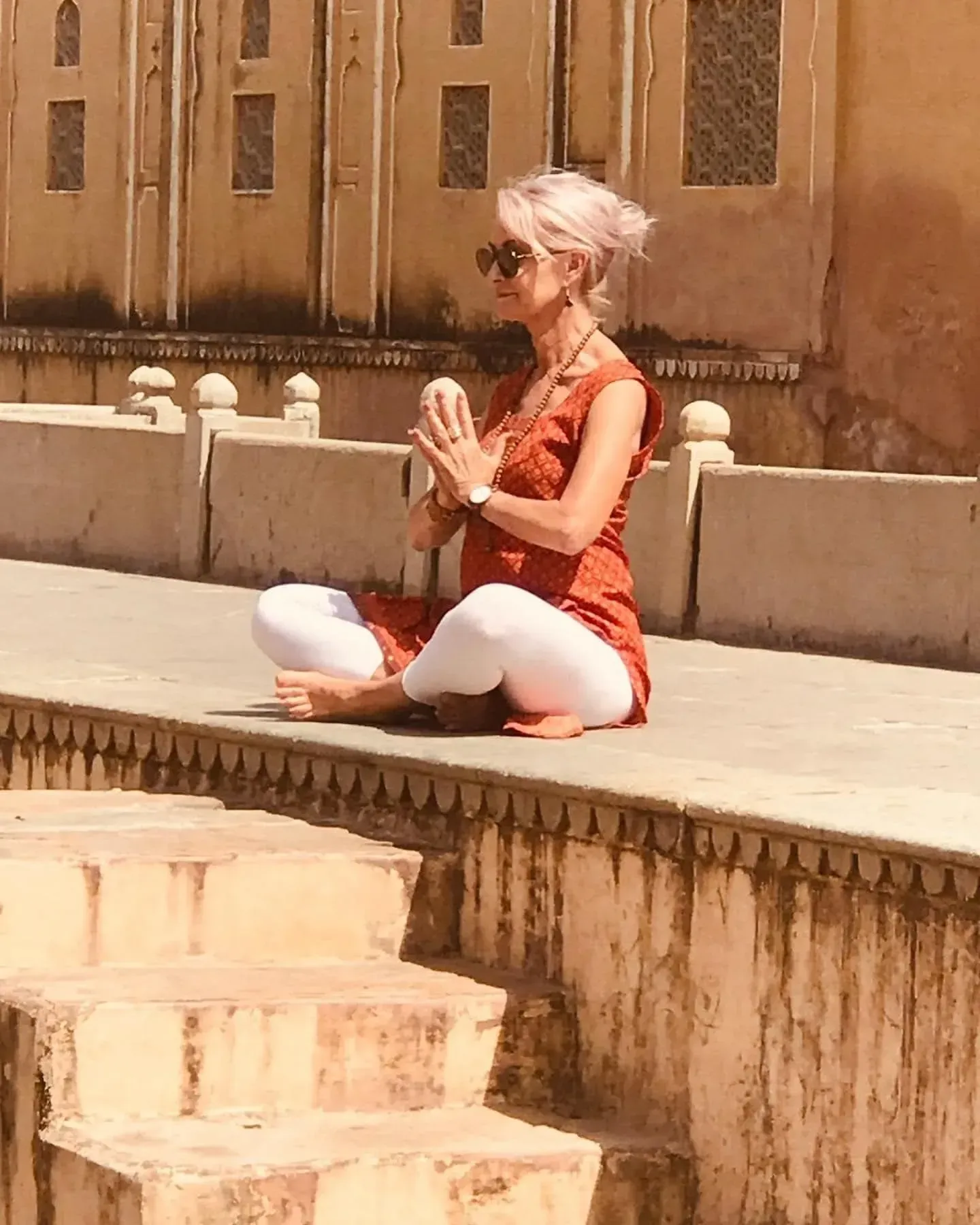 A person in an orange tunic and white pants meditating in a seated pose on the stone edge of a historic building.