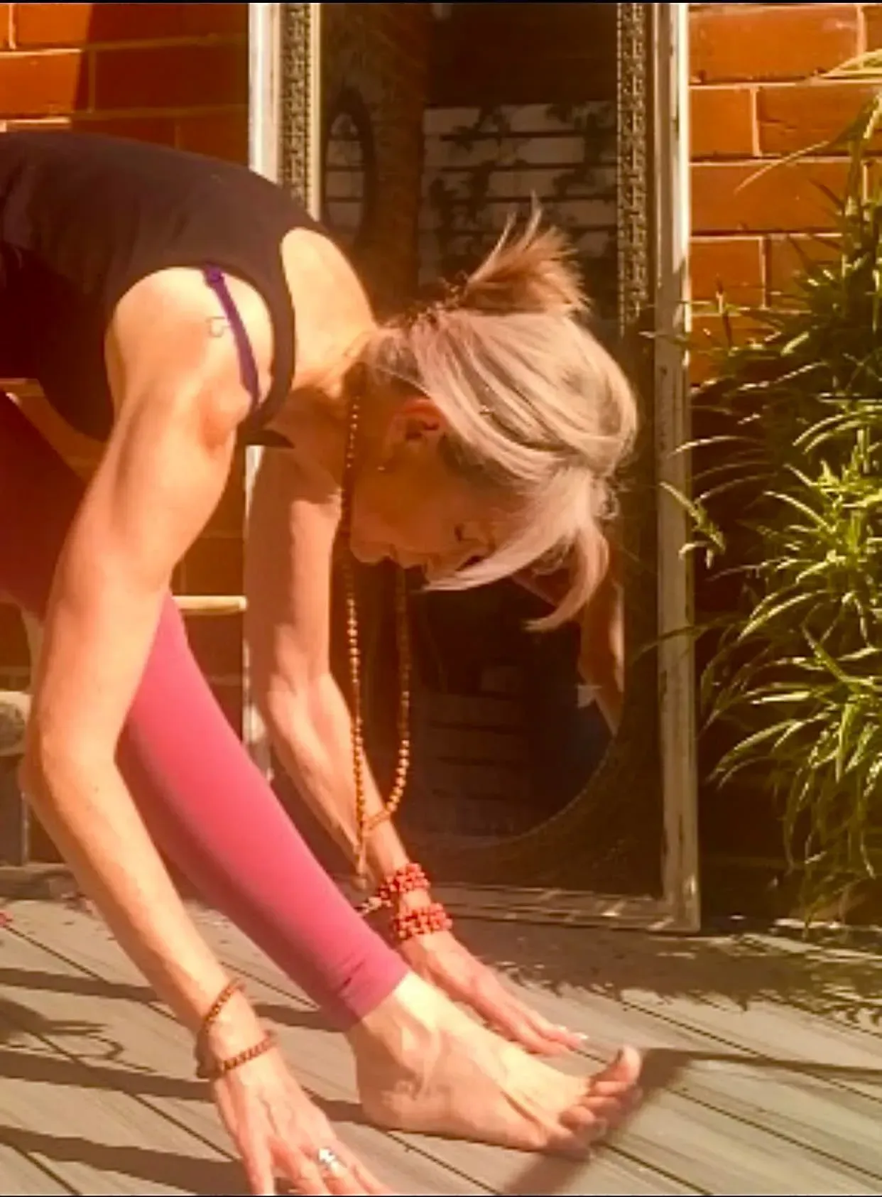 A person in athletic wear performs a forward fold yoga stretch on a wooden deck next to a mirror and a green plant.