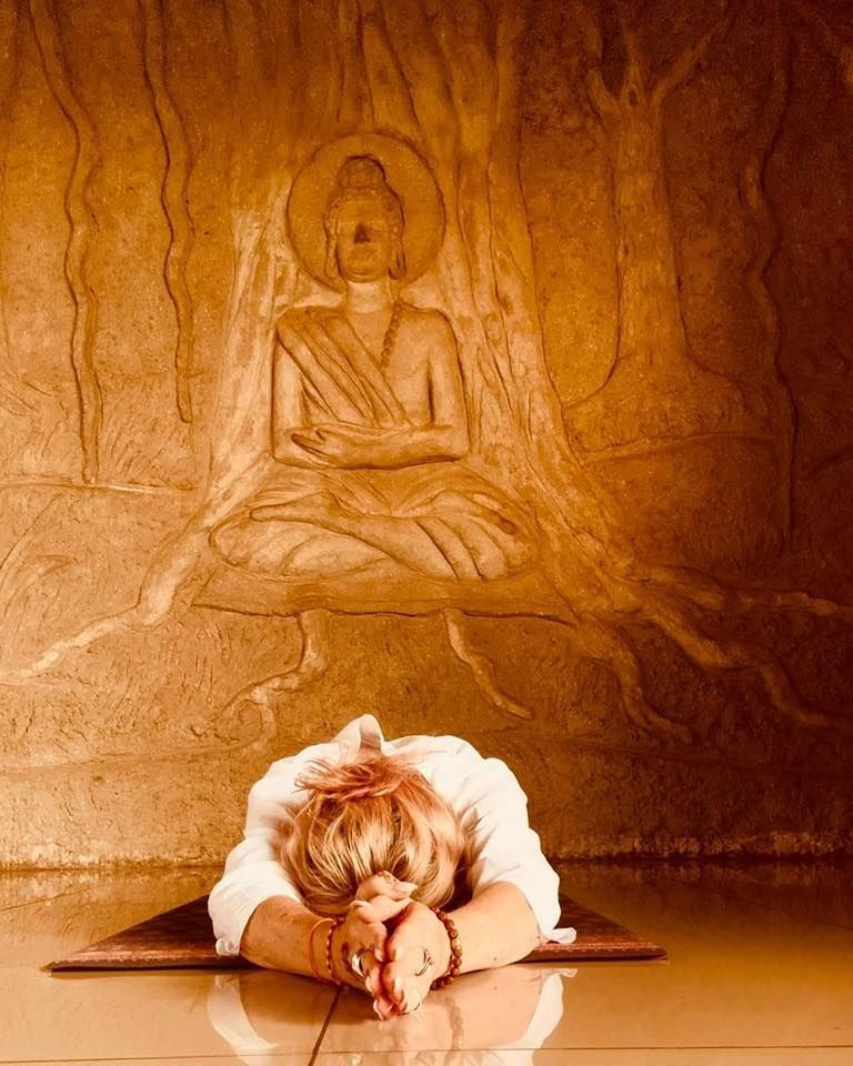 A person in white clothing performs a prostration pose before a textured, golden relief carving of a meditating Buddha.