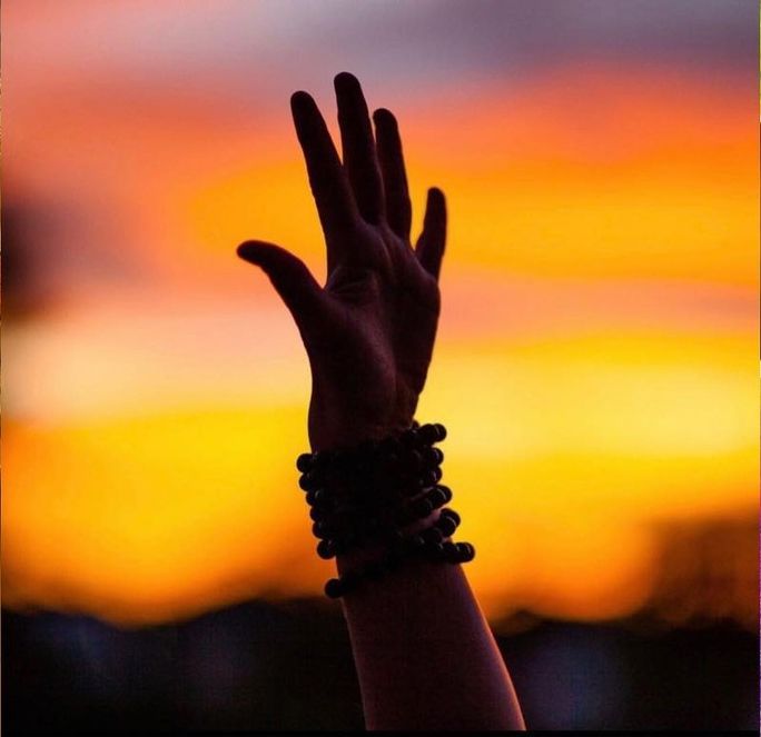 A silhouette of a hand reaching upward against a vibrant, orange and purple sunset background, adorned with beaded rings.