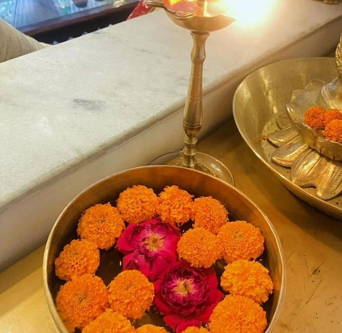 A brass bowl filled with water, marigold flowers, and two pink roses, placed next to a traditional lit brass lamp.
