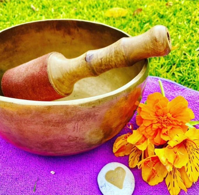 A brass singing bowl with a wooden mallet, bright orange flowers, and a heart-shaped stone on a purple cloth.