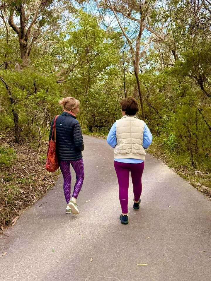 Two people walk side-by-side along a paved trail through a wooded area, wearing casual athletic clothing.