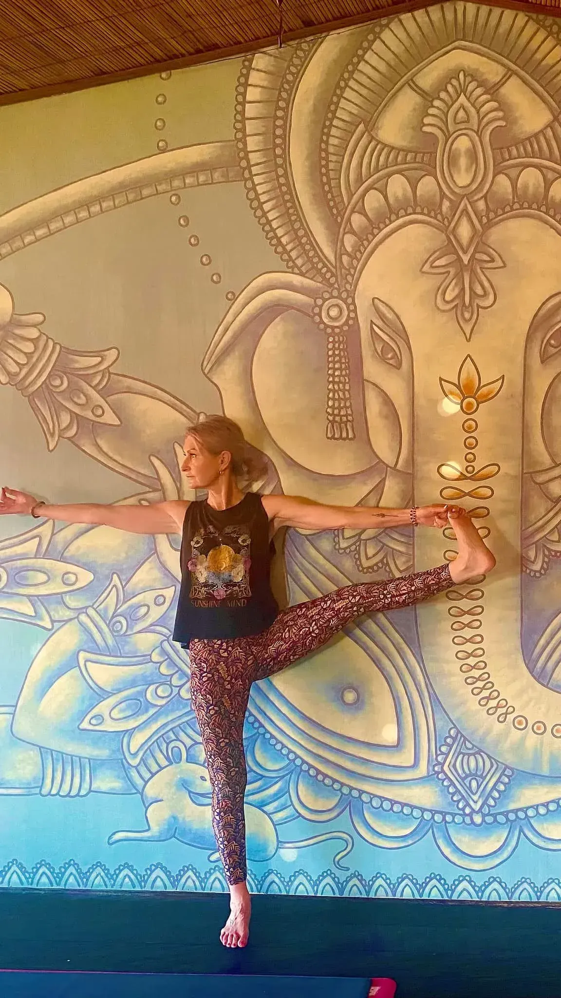 A person performing a yoga pose, balancing on one leg with the other extended, in front of a wall mural of Ganesha.
