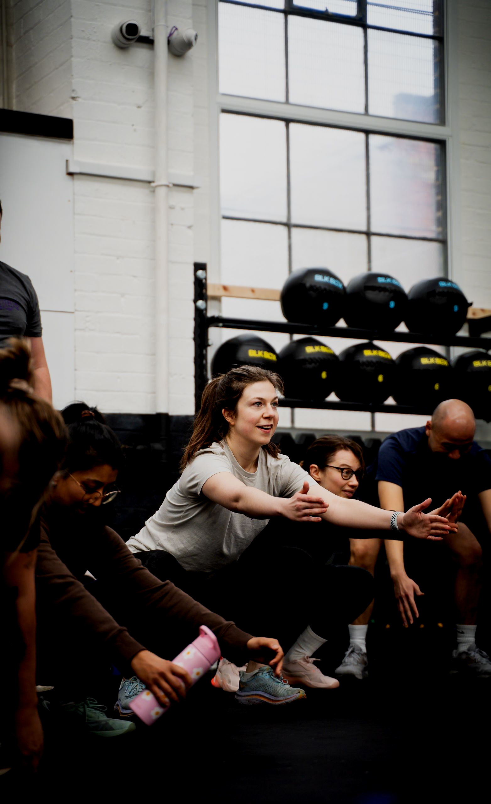 People squatting together in a gym. A woman in the center smiles with arms out. Black exercise balls and windows in the background.