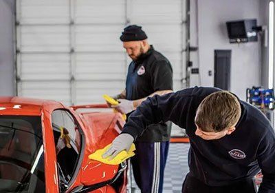 Two men are cleaning a red car in a garage.