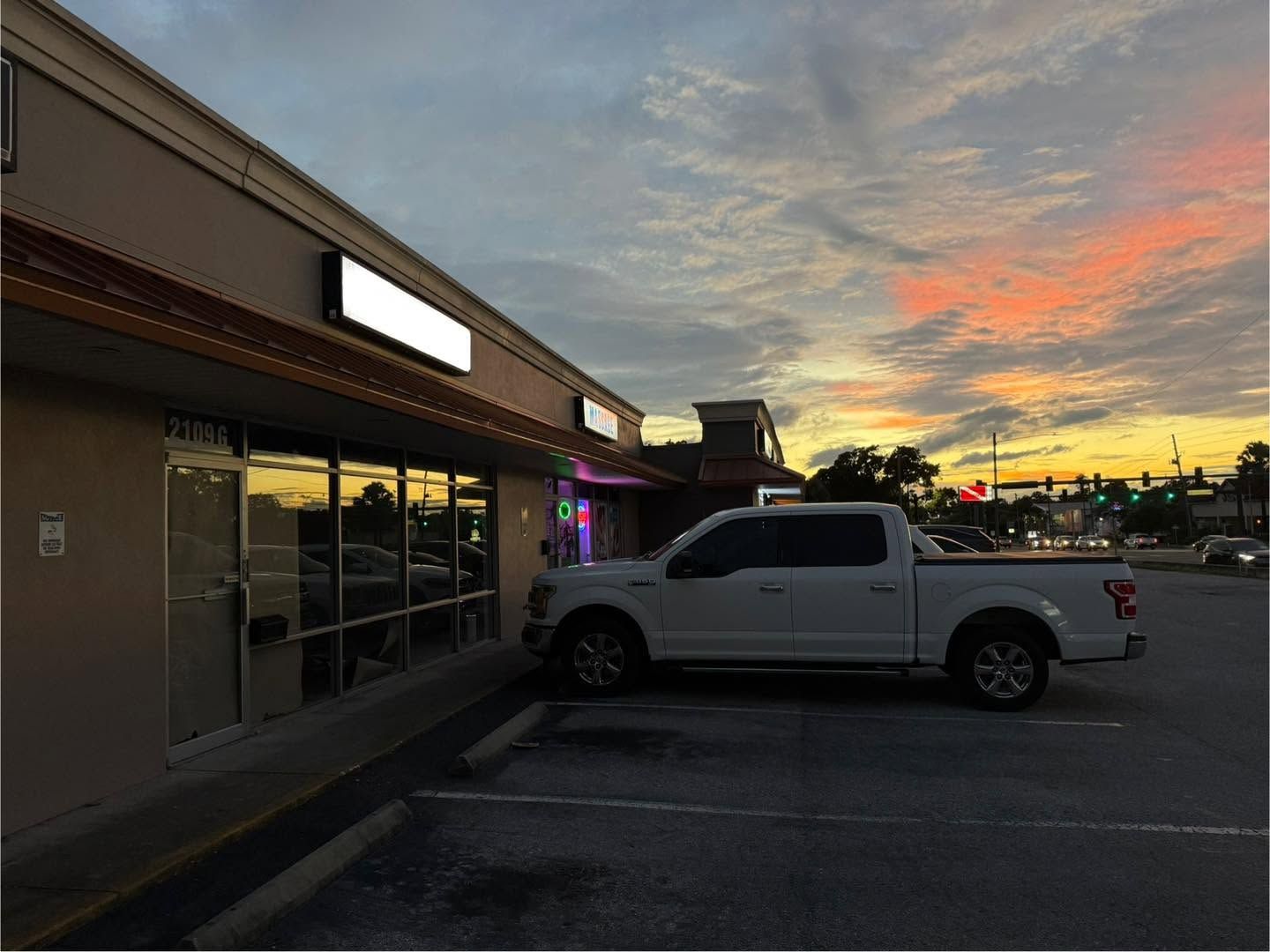 White pickup truck parked outside a storefront at sunset with a colorful orange and pink sky.