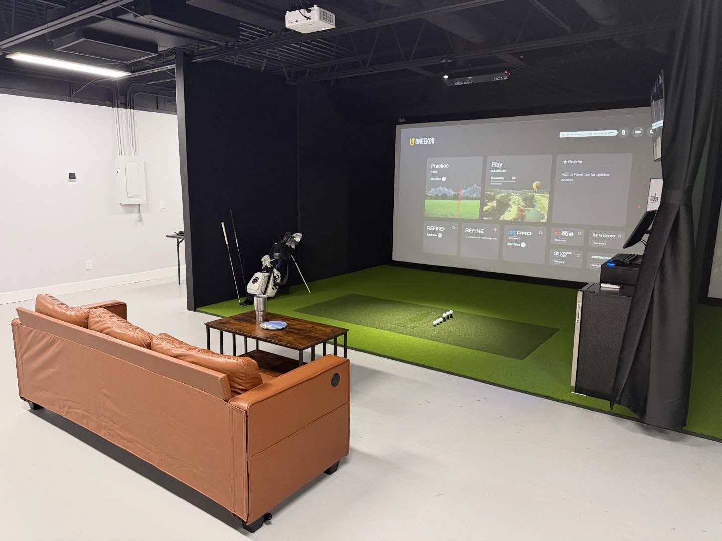 Indoor golf simulator with screen, green turf, and tan couch in a studio space