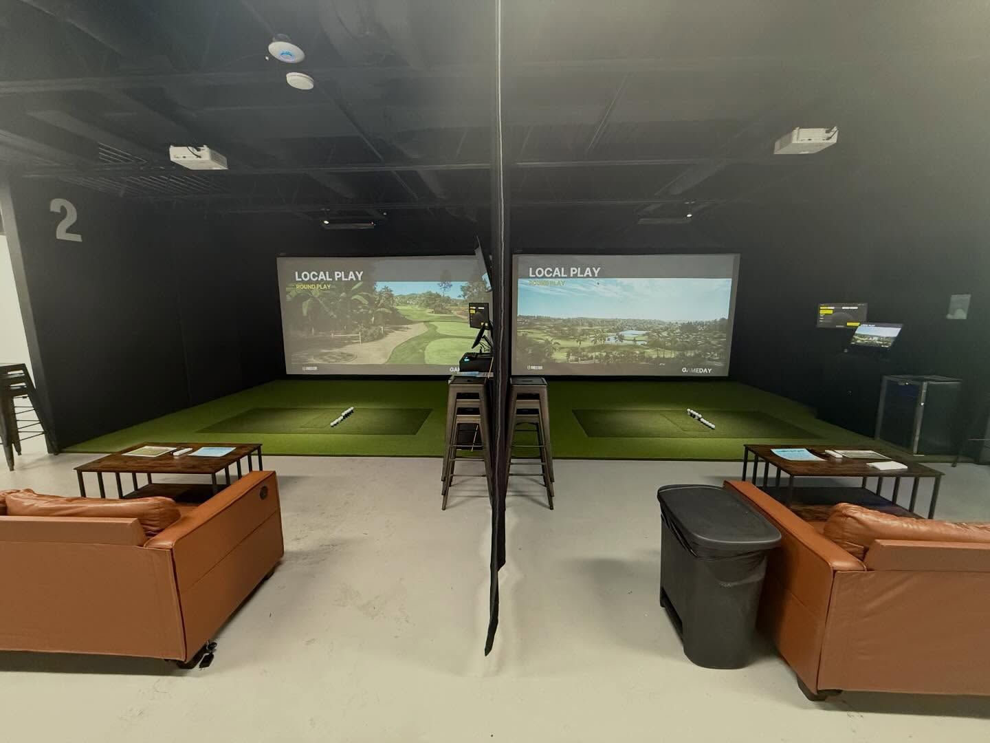 Indoor golf simulator bays with green mats, twin screens, and leather seating in a dark room