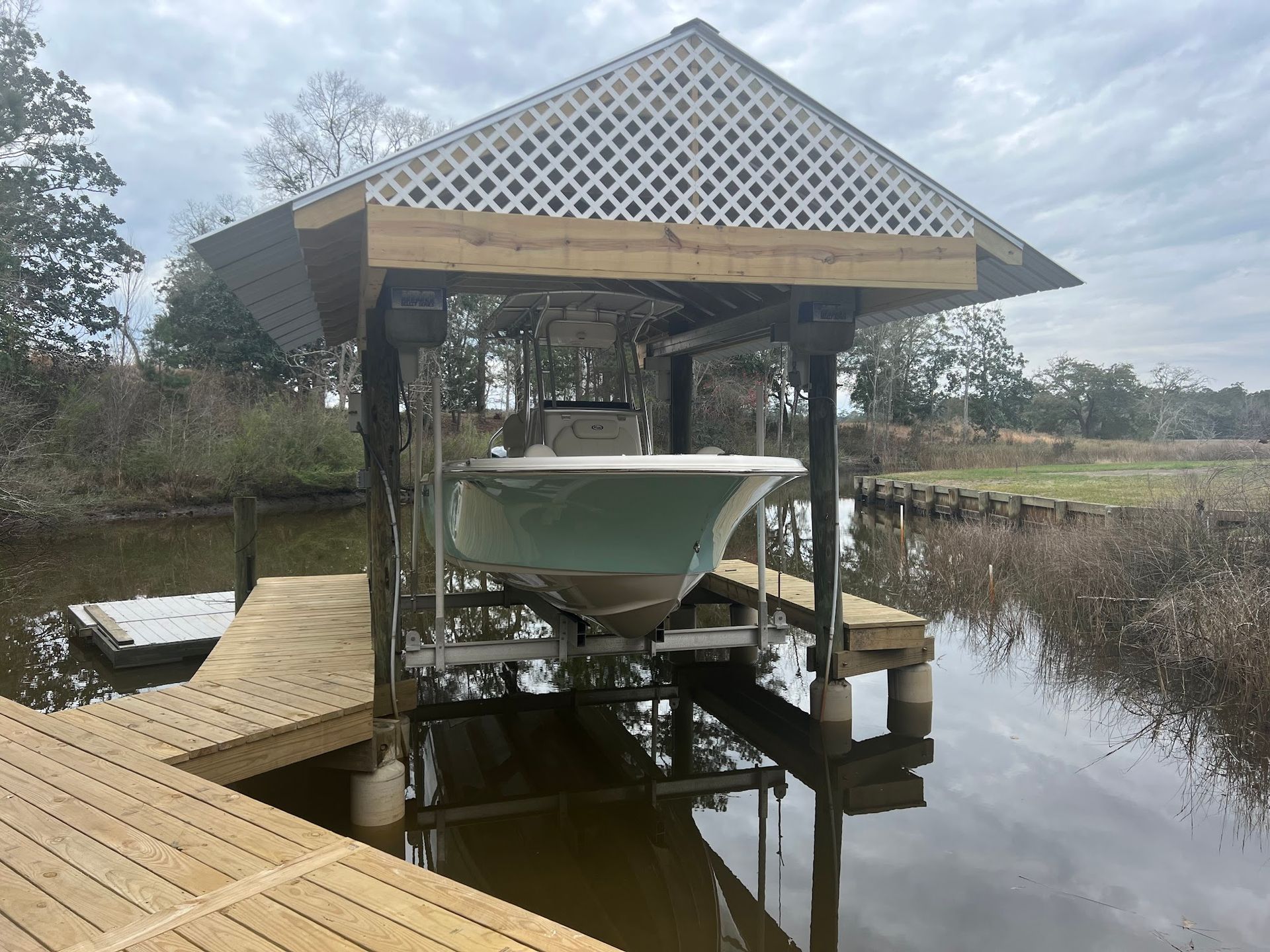 Boat on a lift, covered by a white lattice roof, on a wooden dock over water.