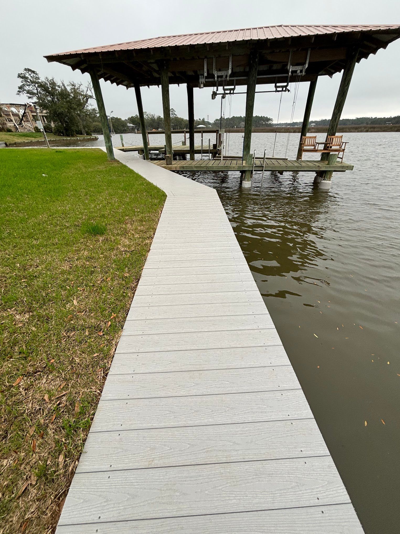 A concrete walkway leads to a covered dock on a lake. The sky is overcast and the grass is green.