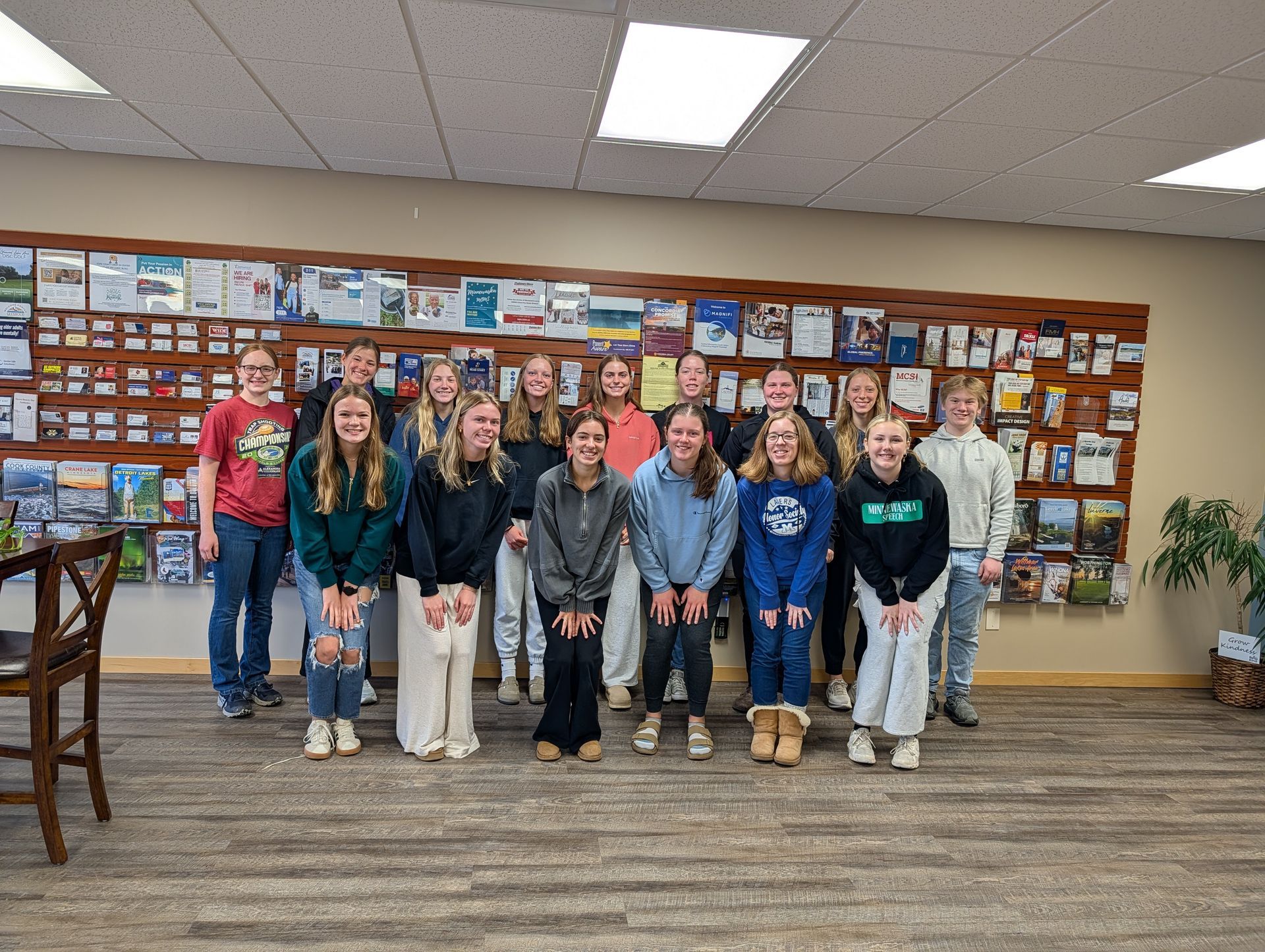 A group of people standing in a row in front of a wall covered in brochures, all smiling toward the camera.