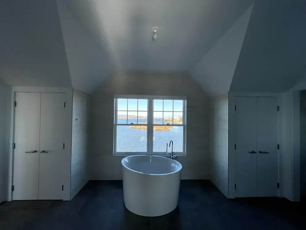 Bathroom with round bathtub in front of a window overlooking a body of water. Two doors flank the window.
