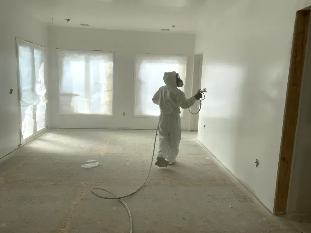 Person in protective suit spraying paint on a white wall in a room; windows covered.