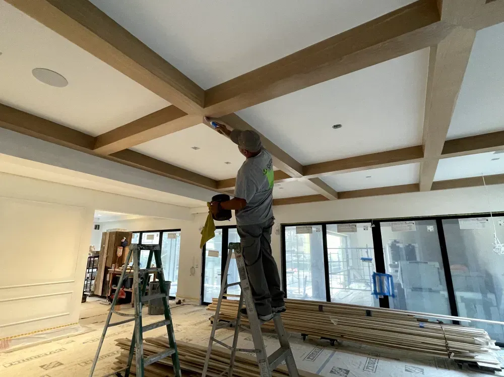 Person painting a ceiling's wooden beams in a room under construction, with a ladder and building materials.